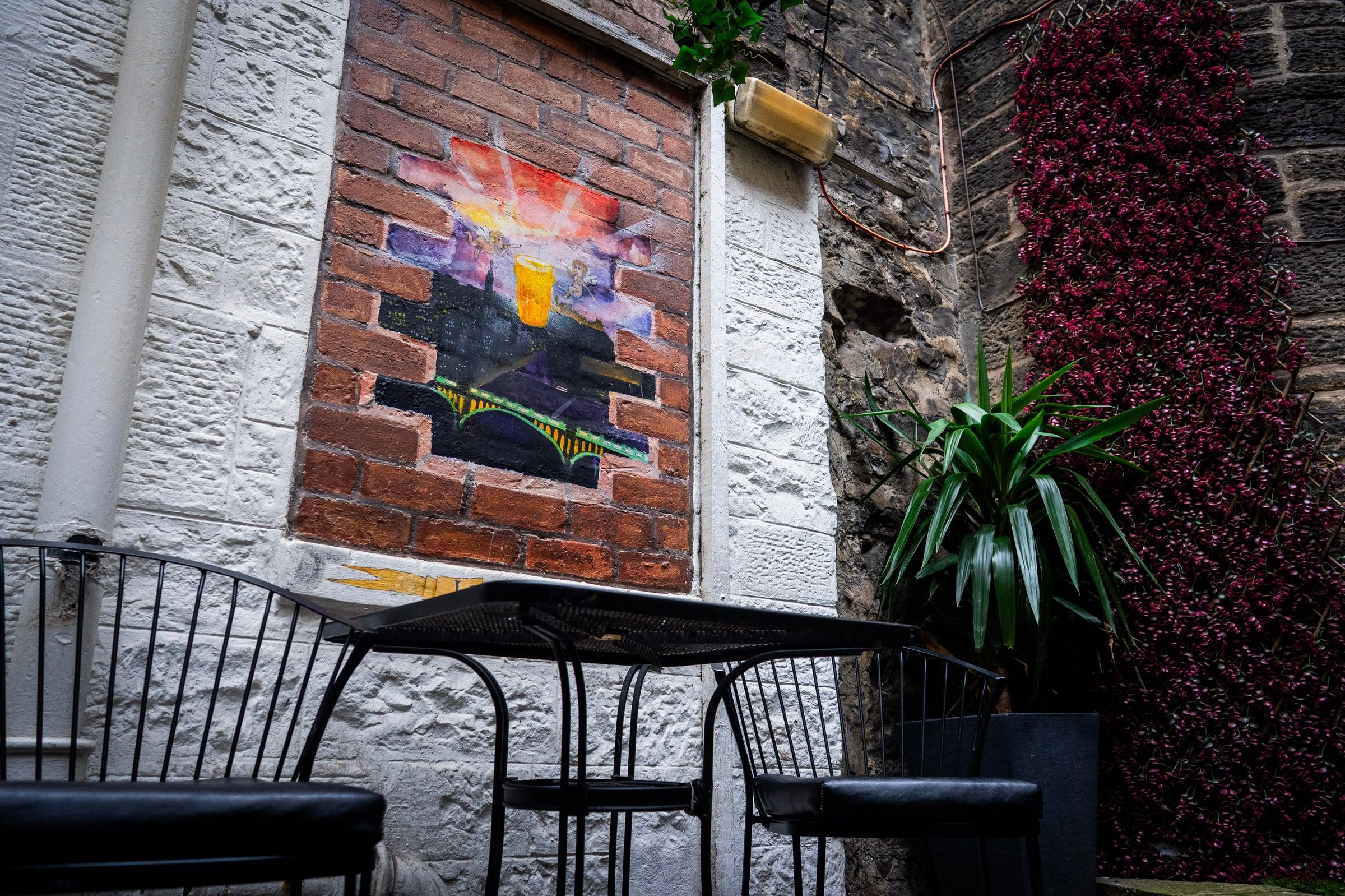 A painted mural of a pint over the city on a brick panel, with black metal chairs and a table in front