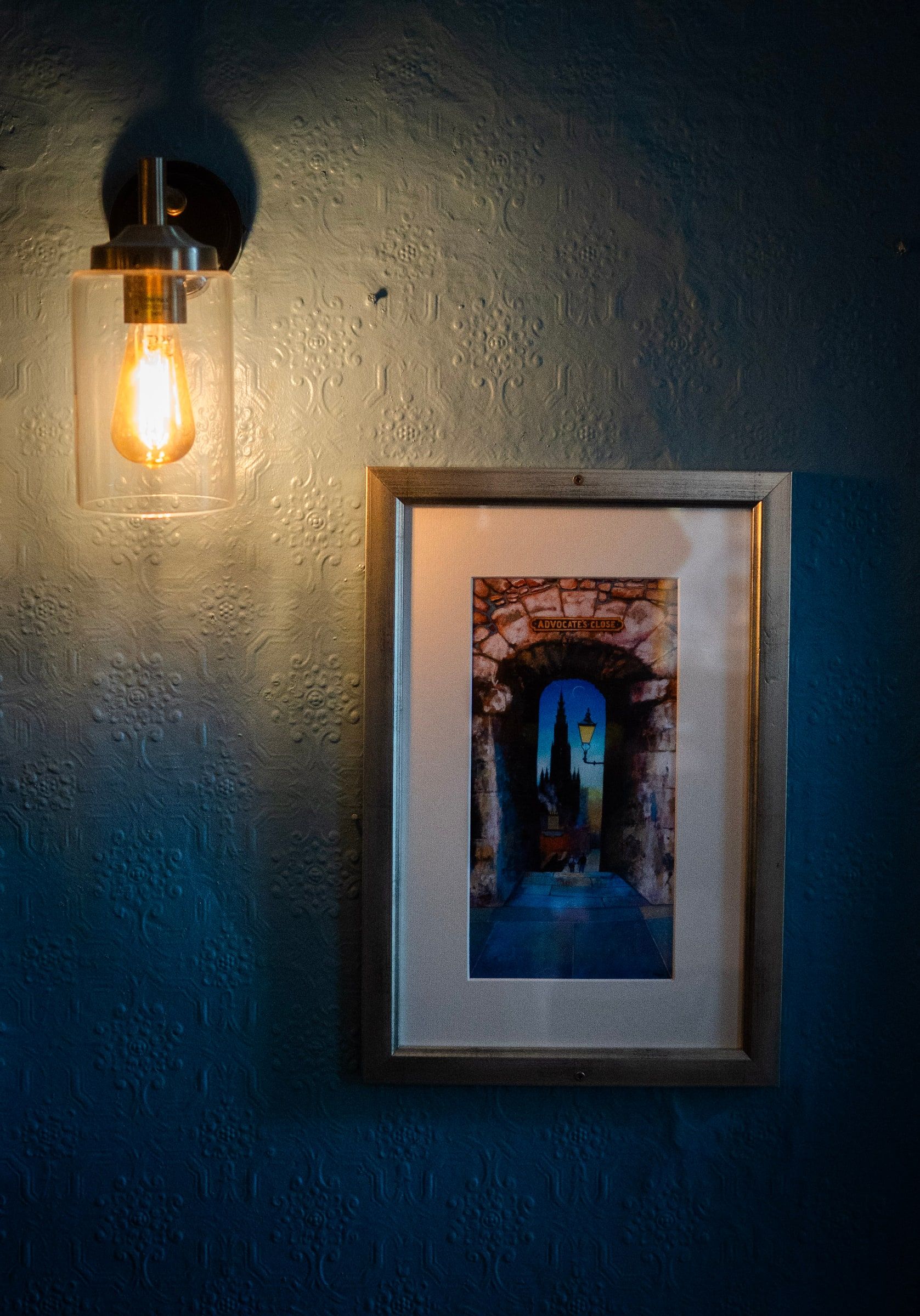 A wall lamp and a framed painting of Advocate’s Close on the blue wall